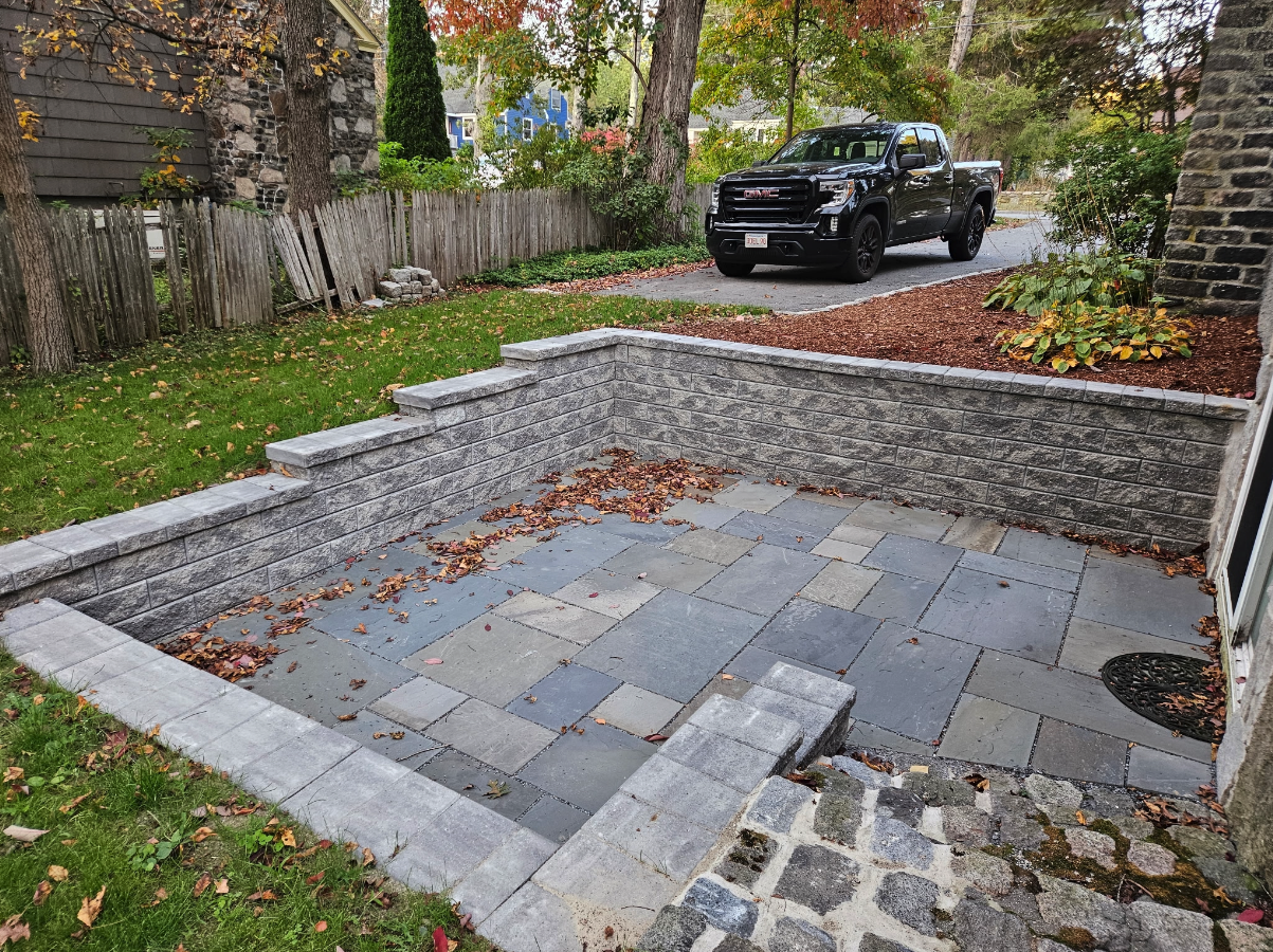 A small stone patio with retaining walls, partially covered in fallen leaves, sits beside a driveway with a black pickup truck parked nearby.