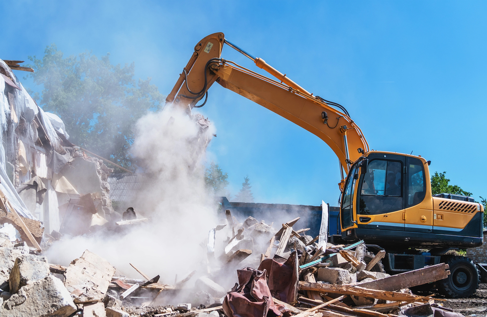 An excavator demolishes a building, surrounded by rubble and dust under a clear blue sky.