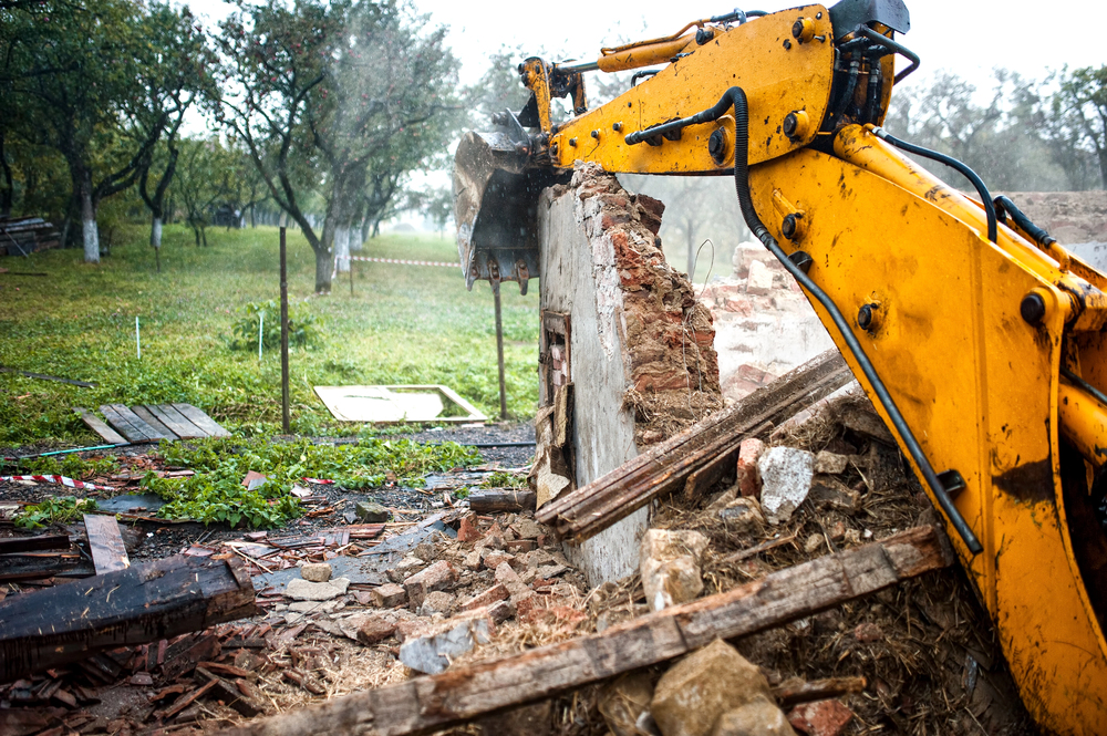 A yellow excavator demolishes a brick wall amidst rubble and debris, with trees and green grass visible in the background.