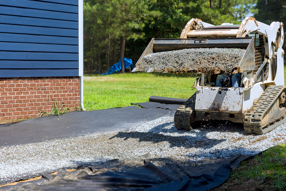 A skid steer loader spreads gravel over black landscaping fabric beside a brick house, preparing for gravel driveway installation and replacement.