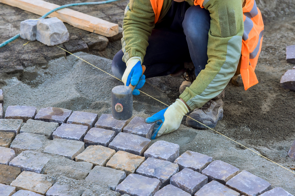 A worker in gloves and a safety vest uses a mallet to install stone pavers in a curved pattern on a sandy base, showcasing the precision often required in gravel driveway installation and replacement at construction sites.