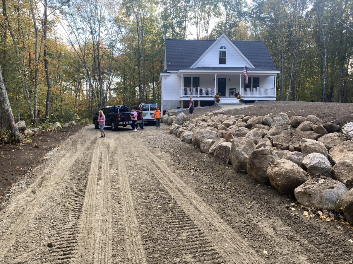 A white house with a front porch sits at the end of a gravel driveway; several people and two vehicles are near the entrance, with large rocks lining part of the yard.