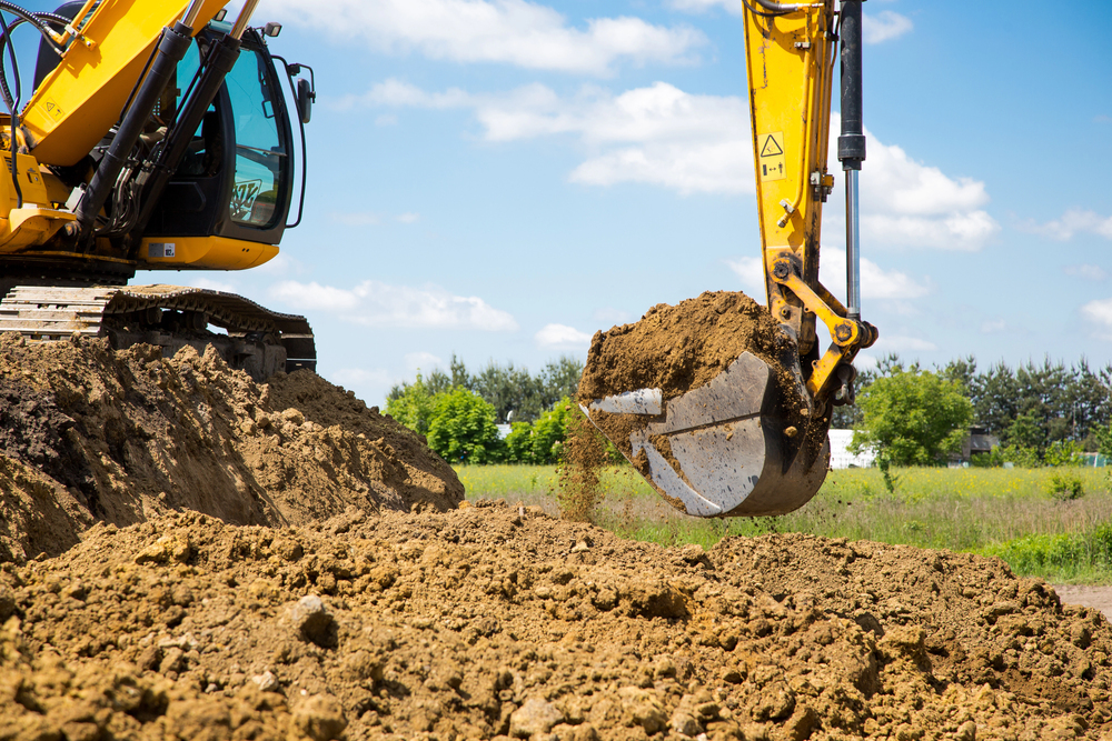A yellow excavator moves a bucketful of soil at a construction site on a sunny day, with green trees and blue sky in the background.