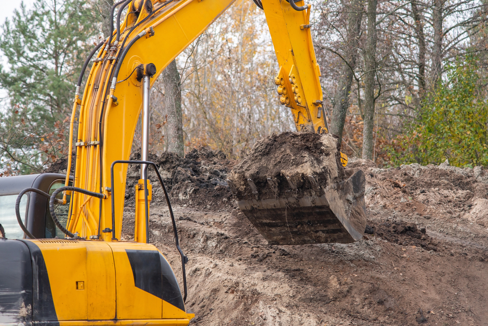 A yellow excavator lifts a bucket of soil at a construction site surrounded by trees and loose dirt.