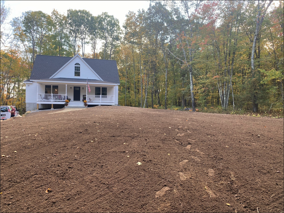 A white house with a front porch sits at the edge of a forested area, with a large, freshly tilled dirt yard in the foreground.