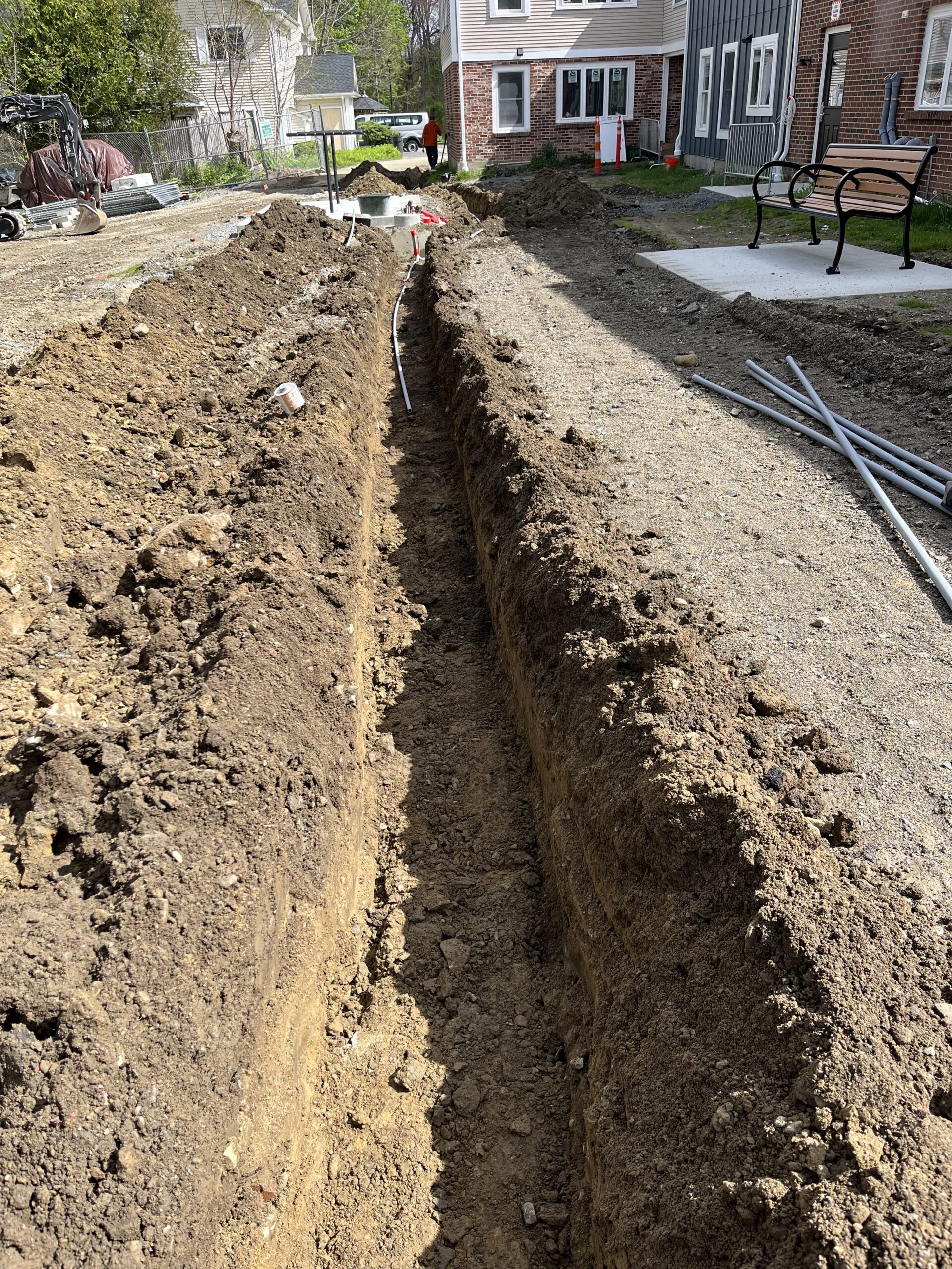 A long, narrow trench is dug in the ground between buildings, with some pipes and construction materials visible along the sides.