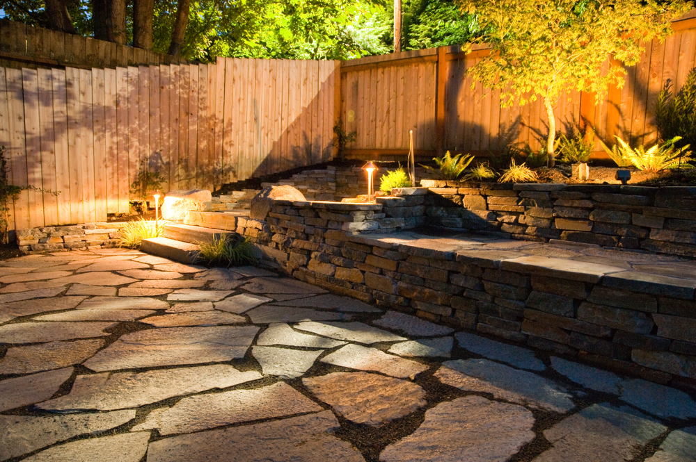 A backyard patio with large stone pavers, terraced stone retaining walls, plants, and warm landscape lighting in the evening.