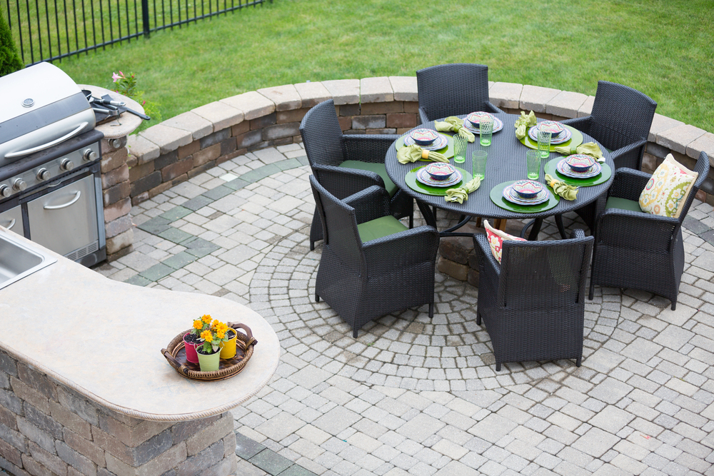 Outdoor patio with a round table set for six, surrounded by wicker chairs, near a built-in grill and countertop with a small flower arrangement. Grass lawn and stone wall in the background.