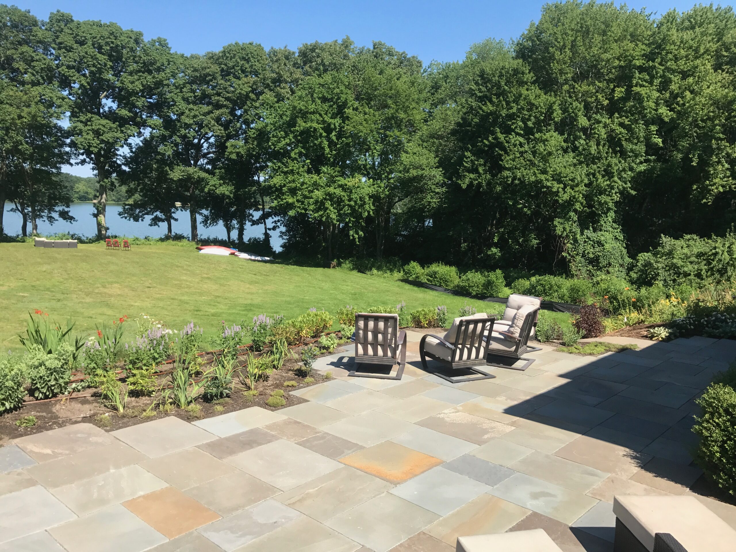 A stone patio with cushioned chairs overlooks a grassy lawn, dense trees, and a lake in the background on a sunny day.