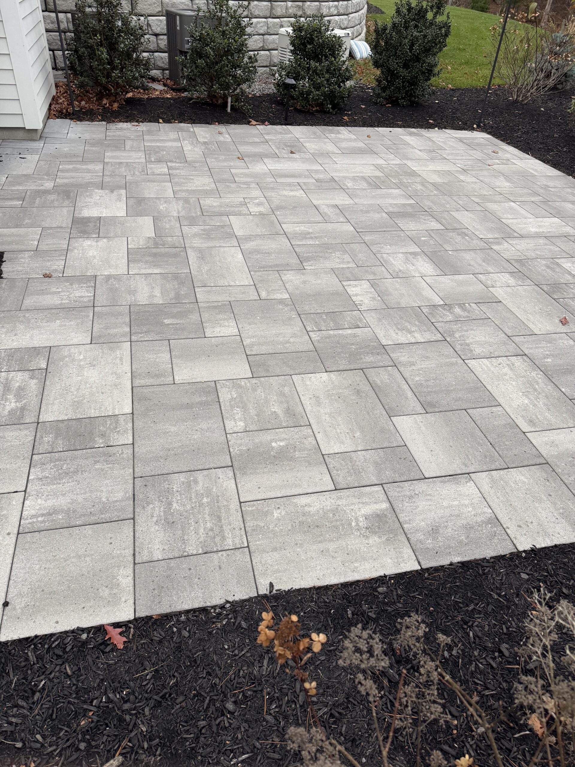 A freshly installed gray stone patio with a geometric pattern, bordered by black mulch, shrubs, and a stone wall in the background.