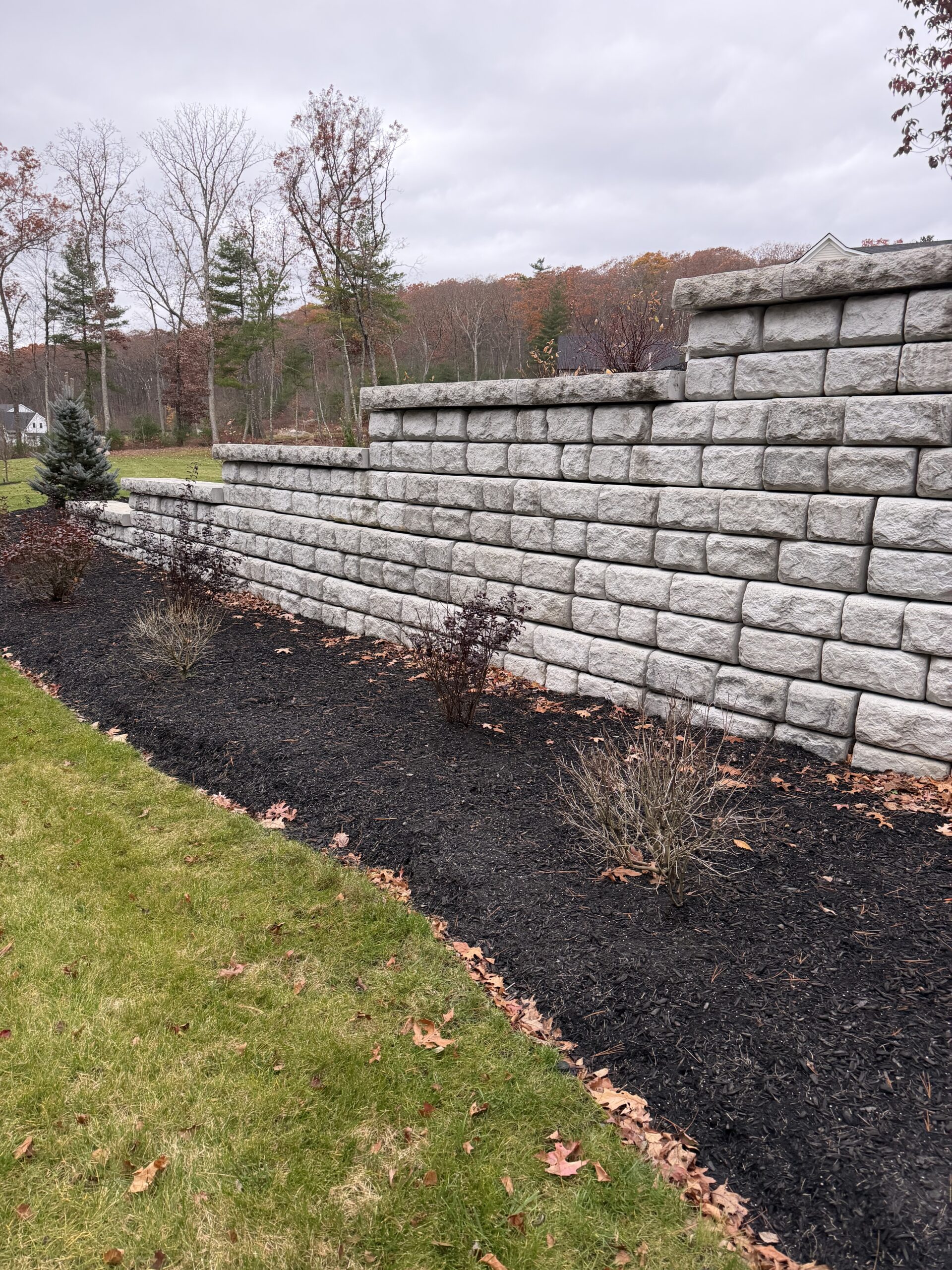 A tiered stone retaining wall borders a landscaped area with mulch and small shrubs; trees and cloudy sky are visible in the background.