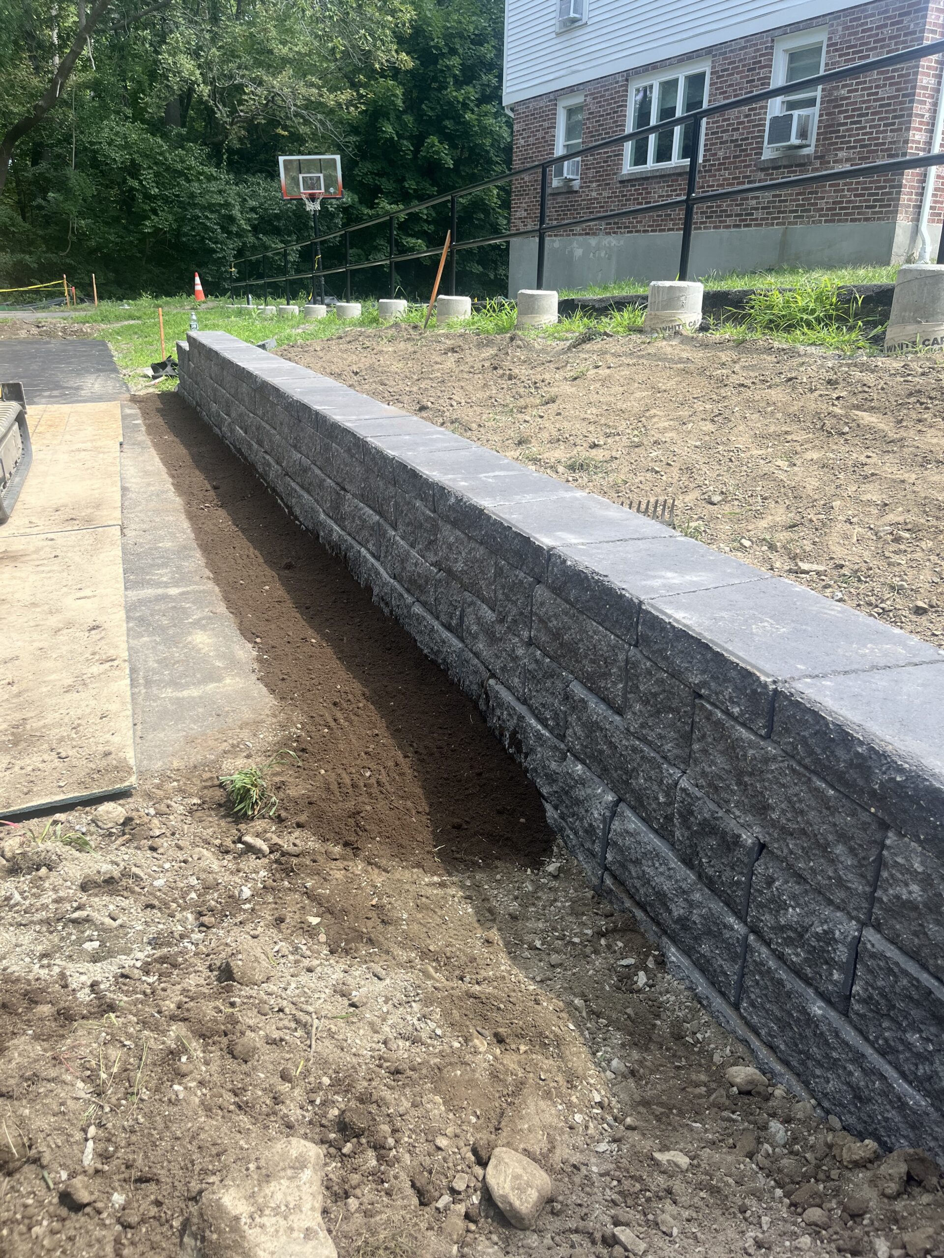 A newly constructed gray stone retaining wall runs alongside a dirt area near a brick building and a basketball hoop in the background.